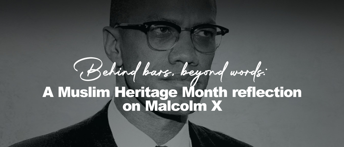 Black-and-white portrait of Malcolm X, shown from the chest up, wearing a dark suit, white shirt, and tie. He looks slightly to the right with a calm, serious expression, his round glasses catching soft light against a textured, light-colored background.
